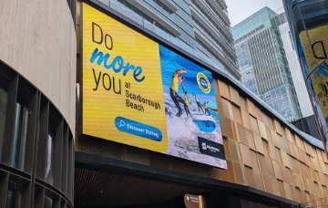 Large outdoor digital billboard mounted on a modern city building displays a bright yellow and blue “Do more you at Scarborough Beach” advertisement with an image of people surfing on ocean waves.